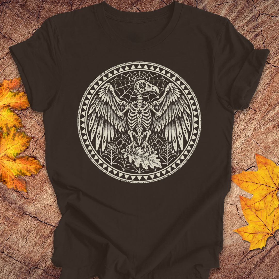 A brown short-sleeve t-shirt with a detailed white circular graphic of a skeletal bird surrounded by webs, laid flat on a rustic wood background with autumn leaves.