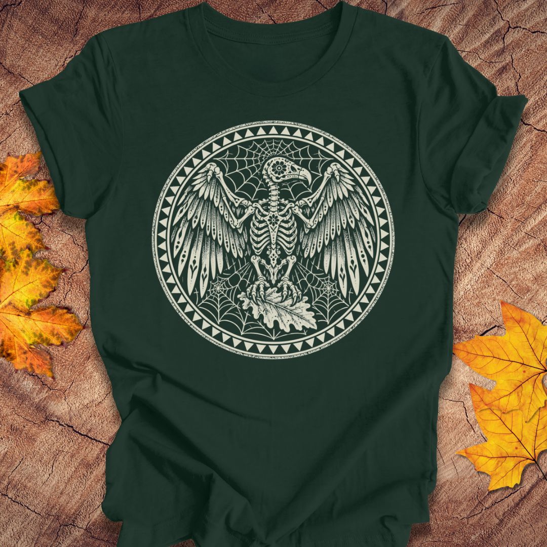 A dark forest green short-sleeve t-shirt with a circular white graphic of a skeletal bird surrounded by webs and geometric borders, laid flat on a rustic wood surface with autumn leaves.