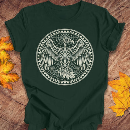 A dark forest green short-sleeve t-shirt with a circular white graphic of a skeletal bird surrounded by webs and geometric borders, laid flat on a rustic wood surface with autumn leaves.