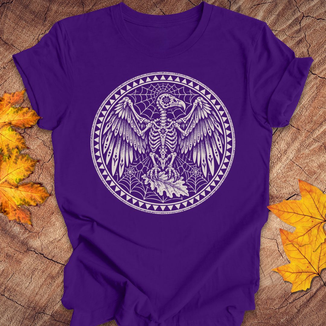 A purple short-sleeve T-shirt laid flat on a wood surface with autumn leaves around it. The shirt has a large circular white graphic on the chest showing a bird skeleton surrounded by webs and decorative borders.