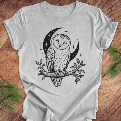 A light gray short-sleeve t-shirt with a black line art graphic of a barn owl perched on a branch under a crescent moon and stars, laid flat on a rustic wood background with pine branches in the corners.