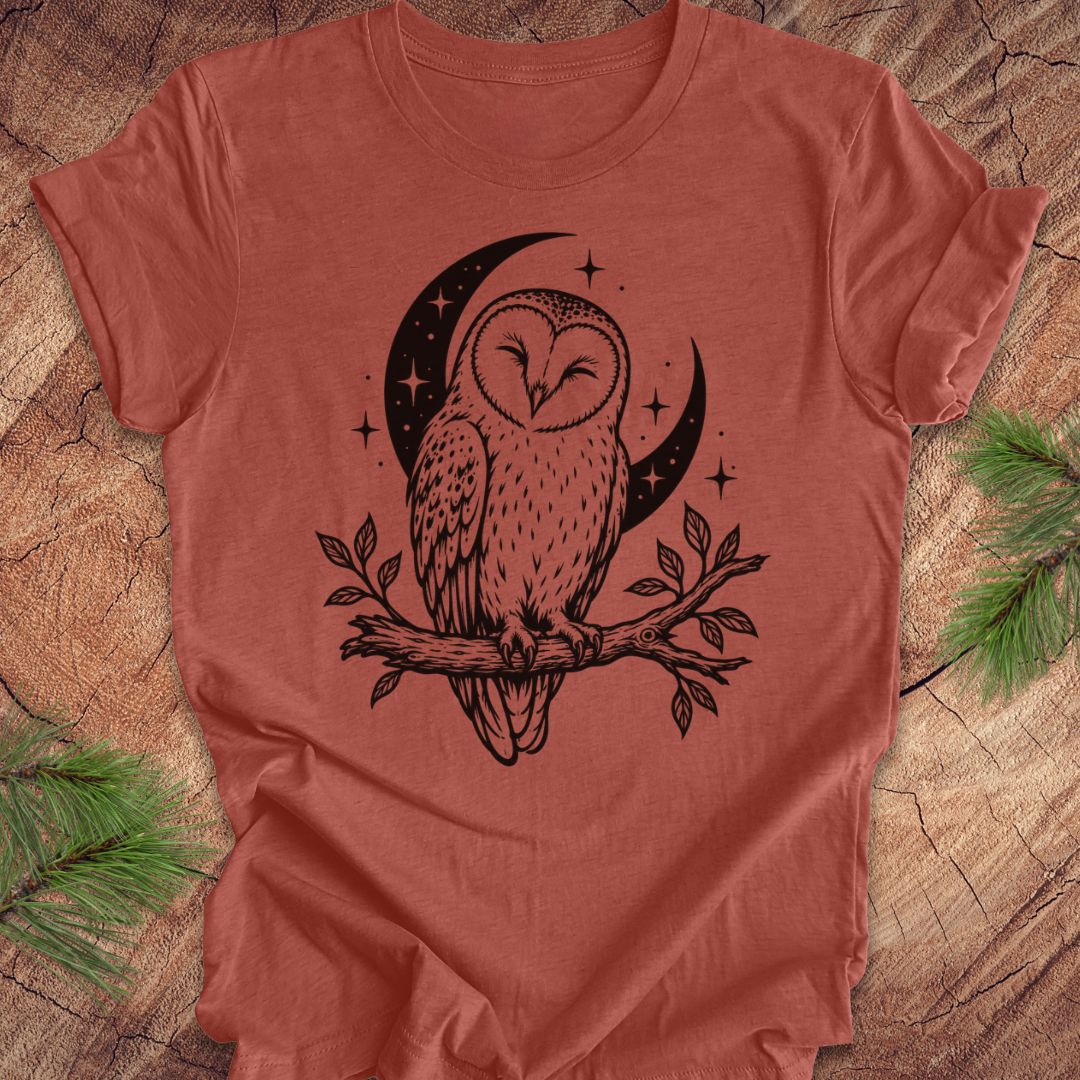 A terracotta-colored short-sleeve t-shirt with a black graphic of an owl perched on a branch in front of a crescent moon and stars, laid flat on a rustic wood background with pine branches in the corners.