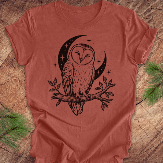 A terracotta-colored short-sleeve t-shirt with a black graphic of an owl perched on a branch in front of a crescent moon and stars, laid flat on a rustic wood background with pine branches in the corners.