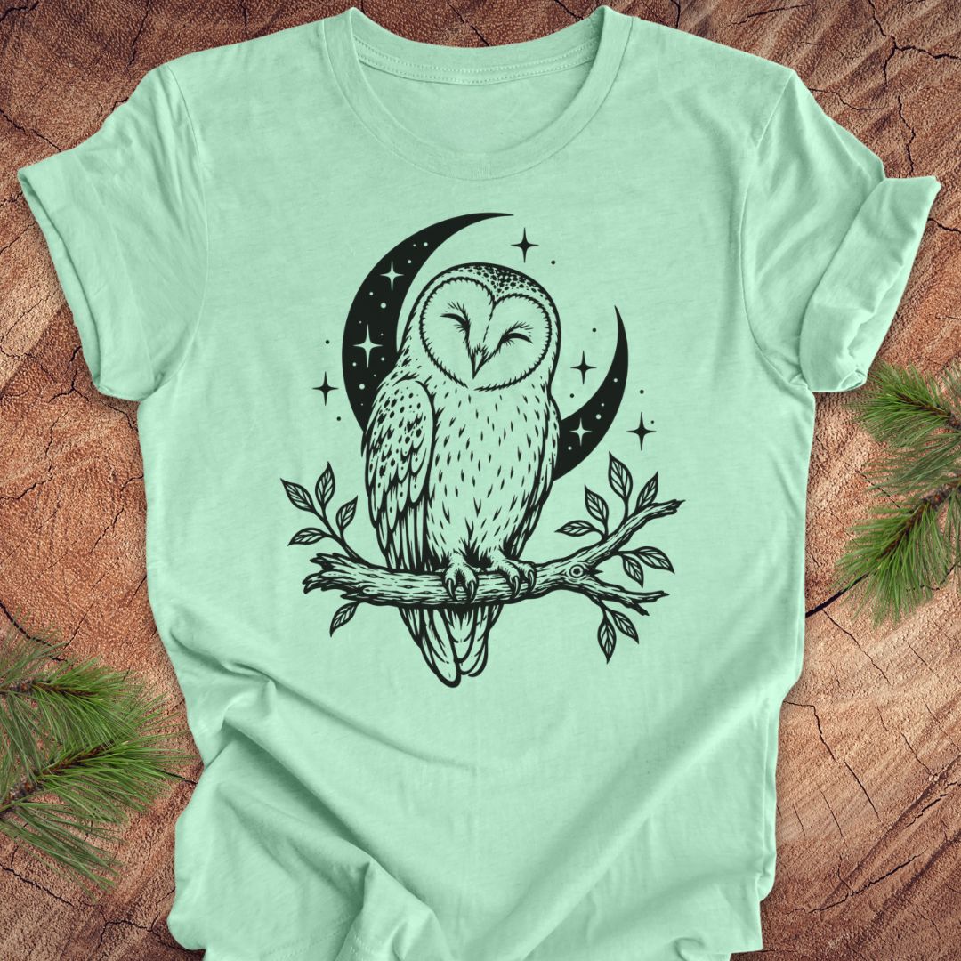 A mint green short-sleeve t-shirt with a black graphic of a barn owl perched on a branch in front of a crescent moon and stars, laid flat on a rustic wood background with pine needles in the corners.