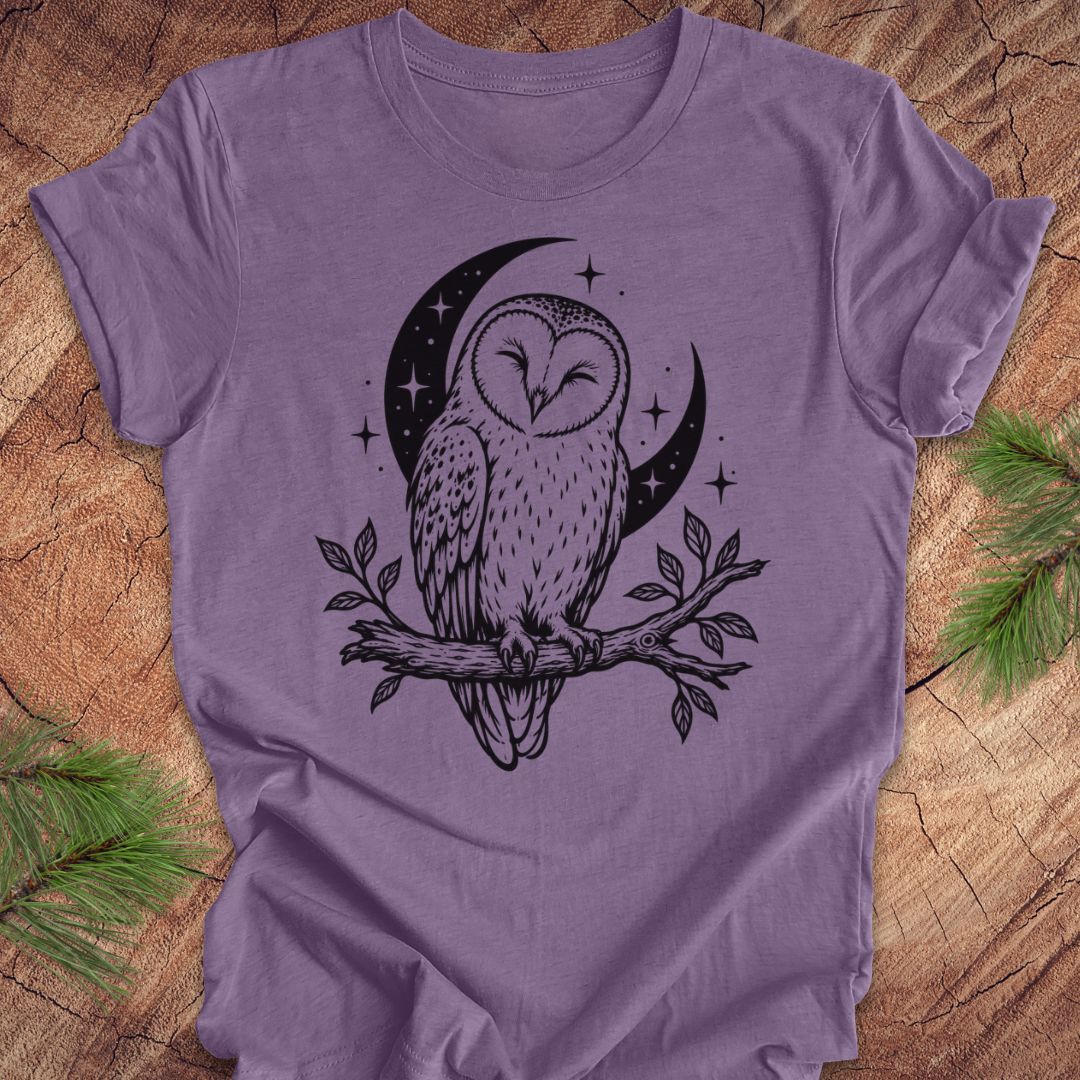 A lavender purple short-sleeve t-shirt with a black graphic of a barn owl perched on a branch in front of a crescent moon and stars, laid flat on a rustic wood background with pine needles in the corners.