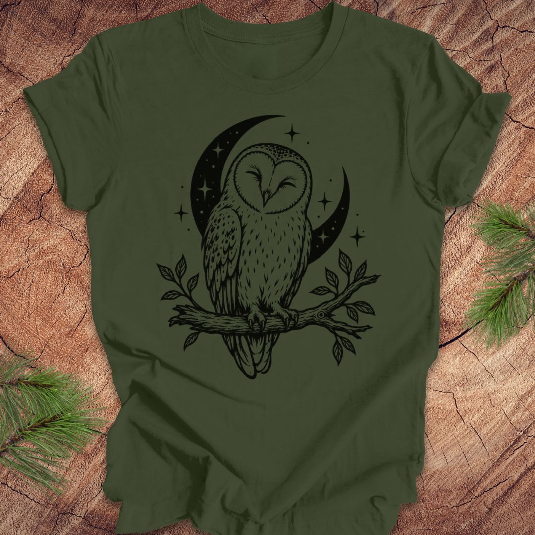 An olive green short-sleeve t-shirt with a black graphic of an owl perched on a branch in front of a crescent moon and stars, laid flat on a rustic wood background with pine branches in the corners.