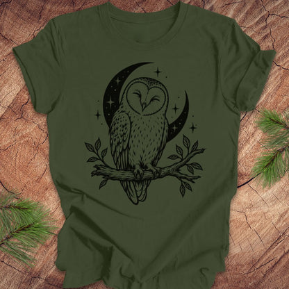 An olive green short-sleeve t-shirt with a black graphic of an owl perched on a branch in front of a crescent moon and stars, laid flat on a rustic wood background with pine branches in the corners.