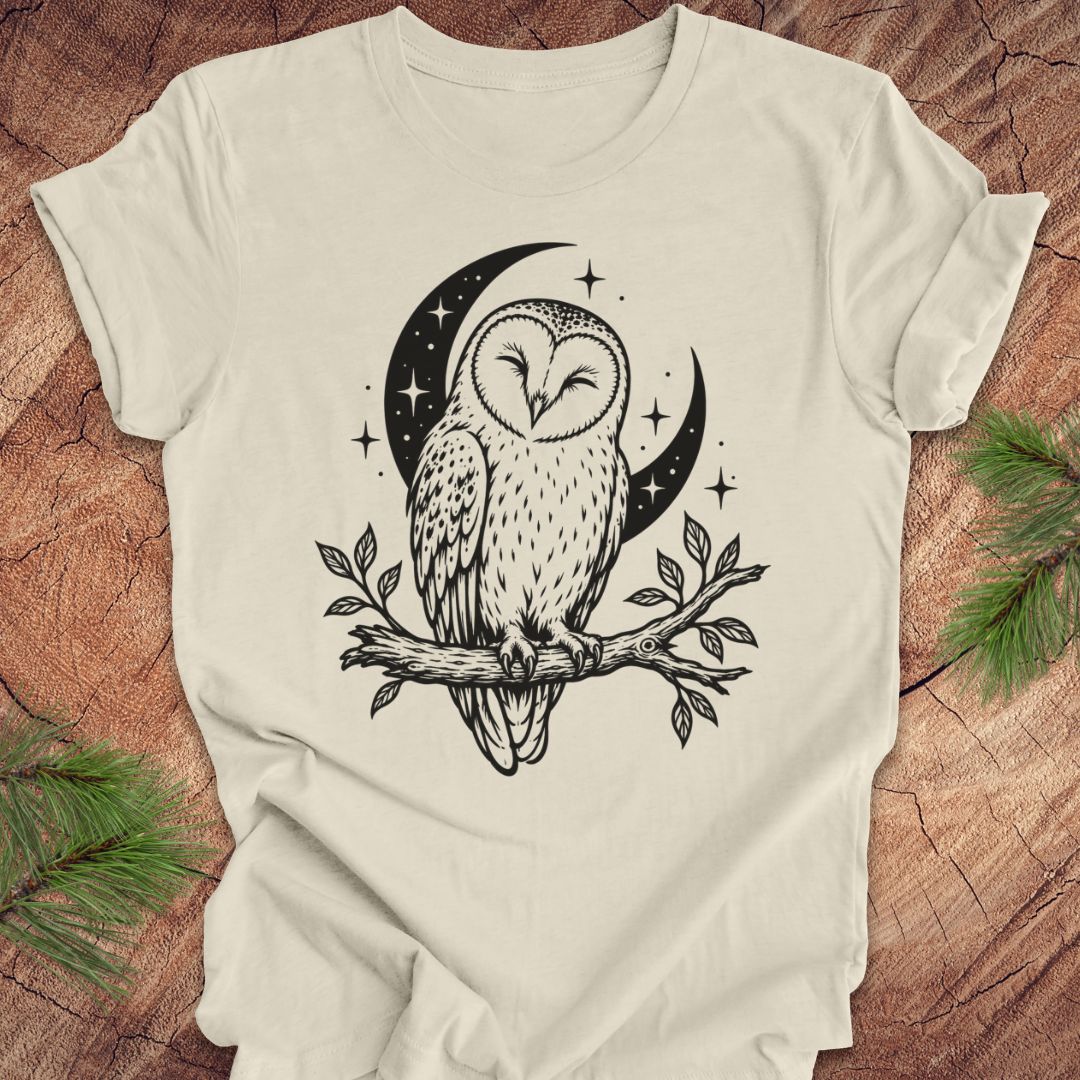 An off-white short-sleeve t-shirt with a black graphic of a barn owl perched on a branch in front of a crescent moon and stars, displayed flat on a rustic wood background with pine needles.