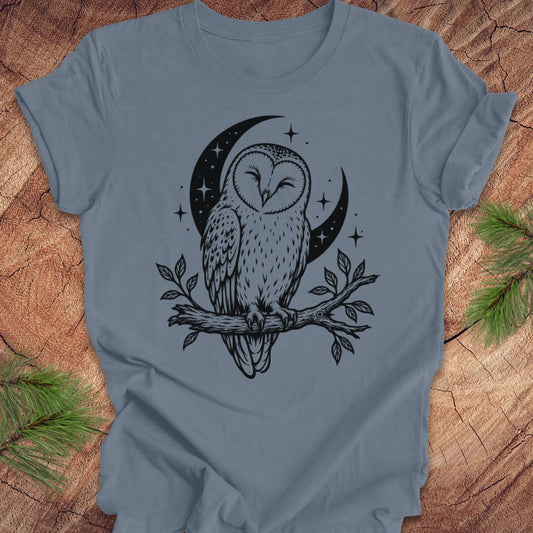 A slate blue short-sleeve t-shirt with a black graphic of an owl perched on a branch in front of a crescent moon and stars, laid flat on a rustic wood background with pine branches in the corners.