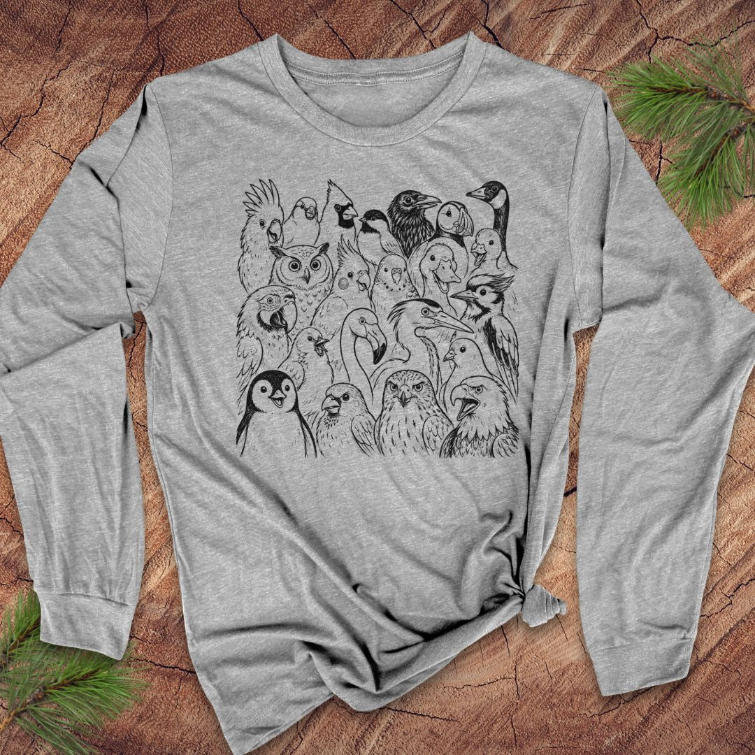 A heather gray long-sleeve t-shirt with a black line art graphic of various birds and owls, styled flat on a rustic wood background with pine needles.