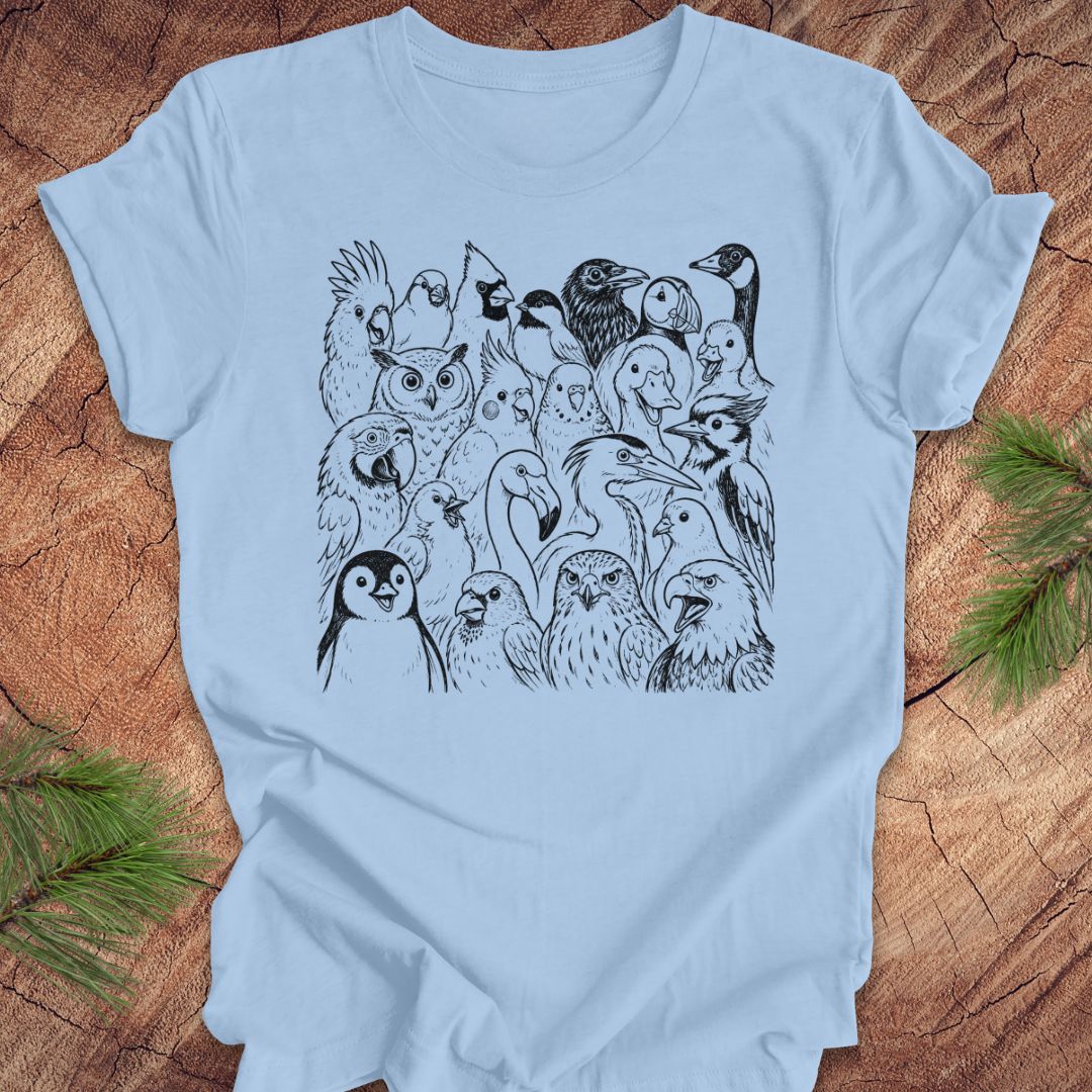 A light blue short-sleeve crewneck t-shirt featuring a black line art graphic of a group of various birds, laid flat on a rustic wood background with pine needles in the corners.