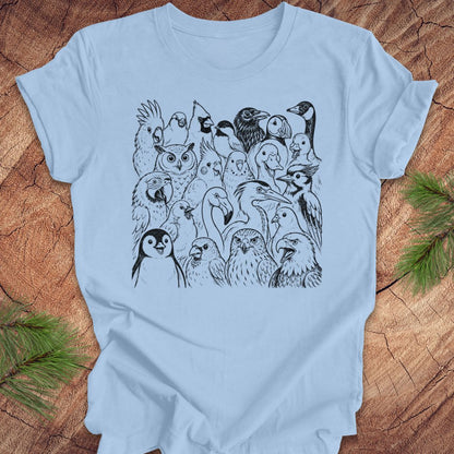 A light blue short-sleeve crewneck t-shirt featuring a black line art graphic of a group of various birds, laid flat on a rustic wood background with pine needles in the corners.