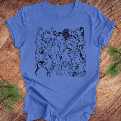 A blue short-sleeve crewneck t-shirt featuring a black line art graphic of a crowd of various birds, laid flat on a rustic wood background with pine needles in the corners.