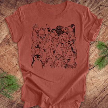 A terracotta-colored short-sleeve t-shirt with a black graphic print of various birds, laid flat on a rustic wood background with pine branches in the corners.