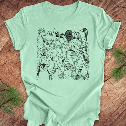 A mint green short-sleeve t-shirt with a black line art graphic of a crowd of various birds, laid flat on a rustic wood background with pine branches in the corners.
