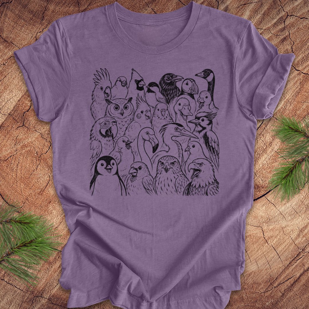 A lavender purple short-sleeve t-shirt with a black line art graphic of various birds, laid flat on a rustic wood background with pine needles in the corners.