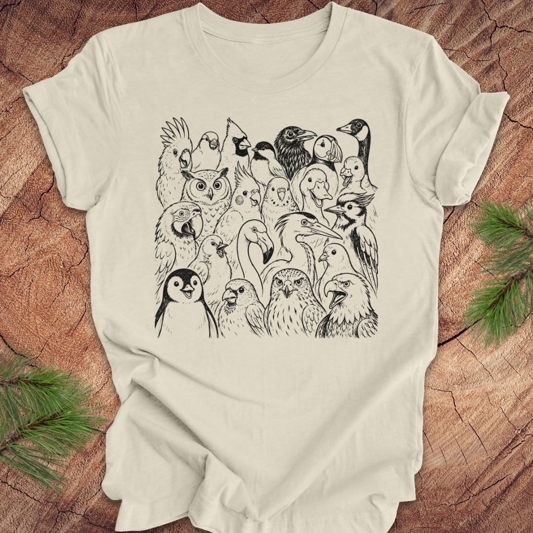 An beige short-sleeve t-shirt with a detailed black line art graphic of various birds and owls, laid flat on a rustic wood background with pine branches in the corners.