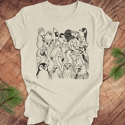 An beige short-sleeve t-shirt with a detailed black line art graphic of various birds and owls, laid flat on a rustic wood background with pine branches in the corners.