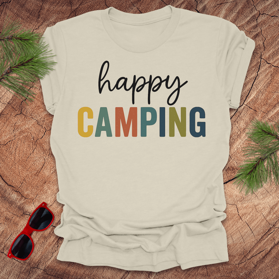 Beige t-shirt with 'happy camping' text on a wooden surface with sunglasses and pine branches.