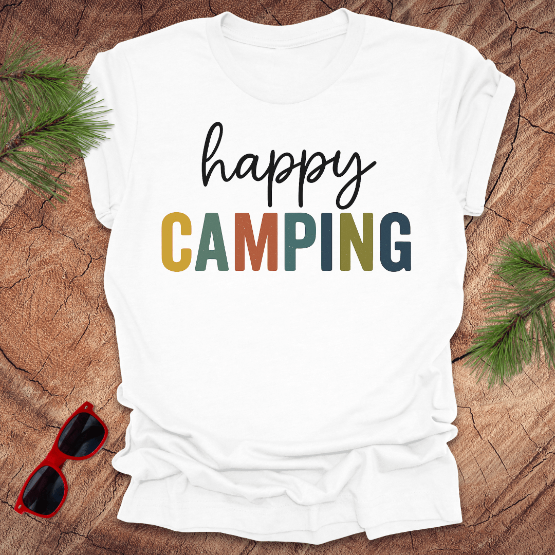 White t-shirt with 'happy CAMPING' text on a wooden surface with sunglasses and pine branches.