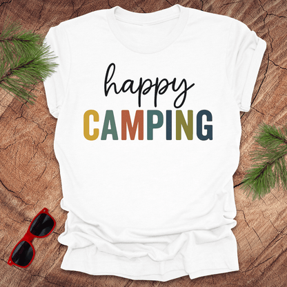White t-shirt with 'happy CAMPING' text on a wooden surface with sunglasses and pine branches.