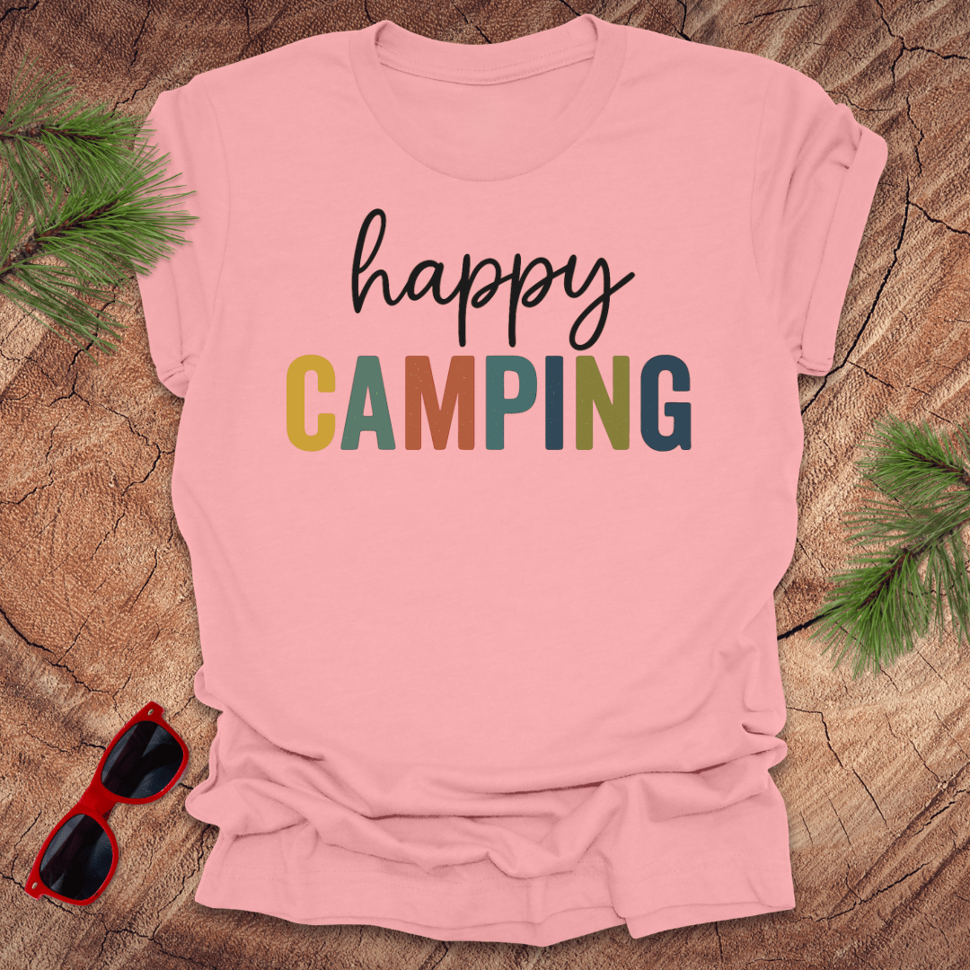 Pink t-shirt with 'happy Camping' text on a wooden surface with sunglasses and pine branches.