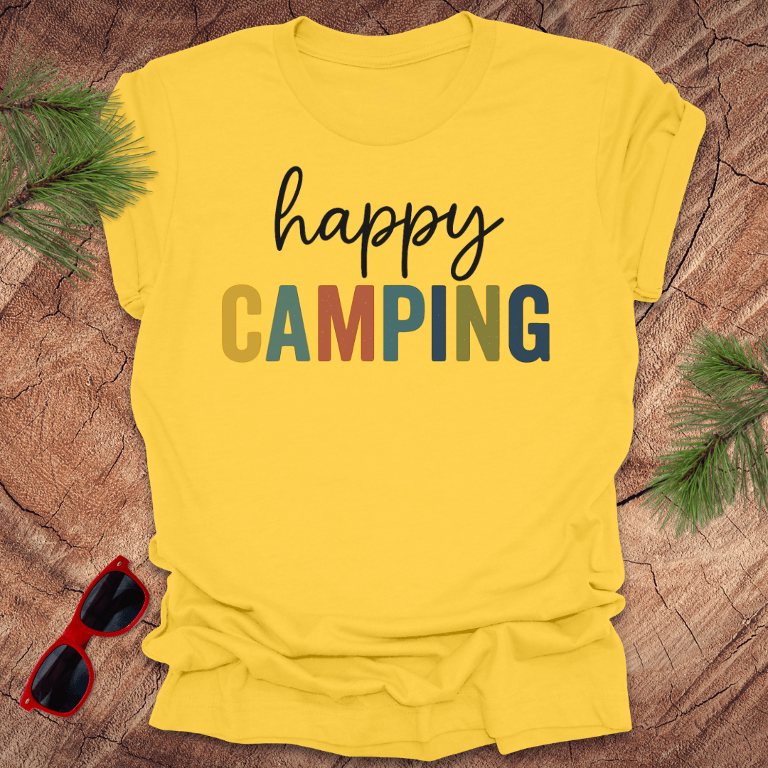 Yellow t-shirt with 'happy Camping' text on a wooden surface with sunglasses and greenery.