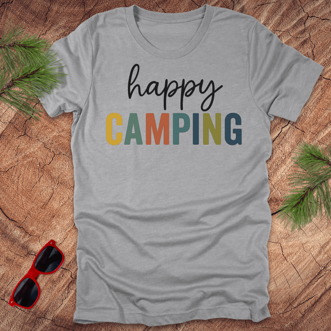 Gray t-shirt with 'happy CAMPING' text on a wooden surface with sunglasses and pine branches.