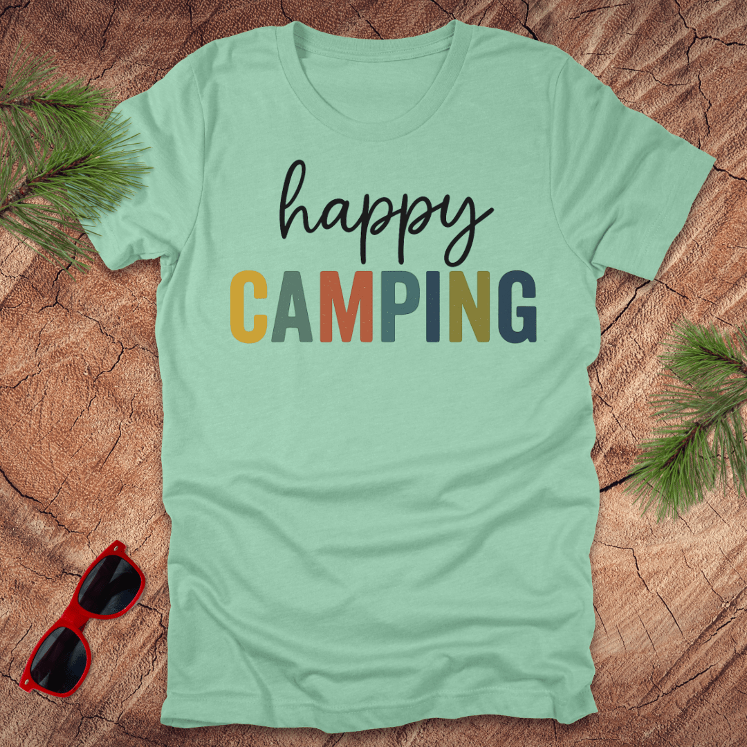 Mint green t-shirt with 'happy CAMPING' text on a wooden surface with sunglasses and pine branches.