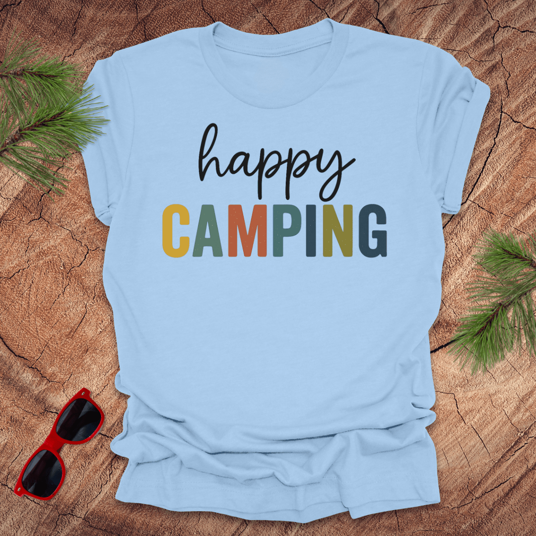 Light blue t-shirt with 'happy Camping' text on a wooden surface with sunglasses and pine branches.