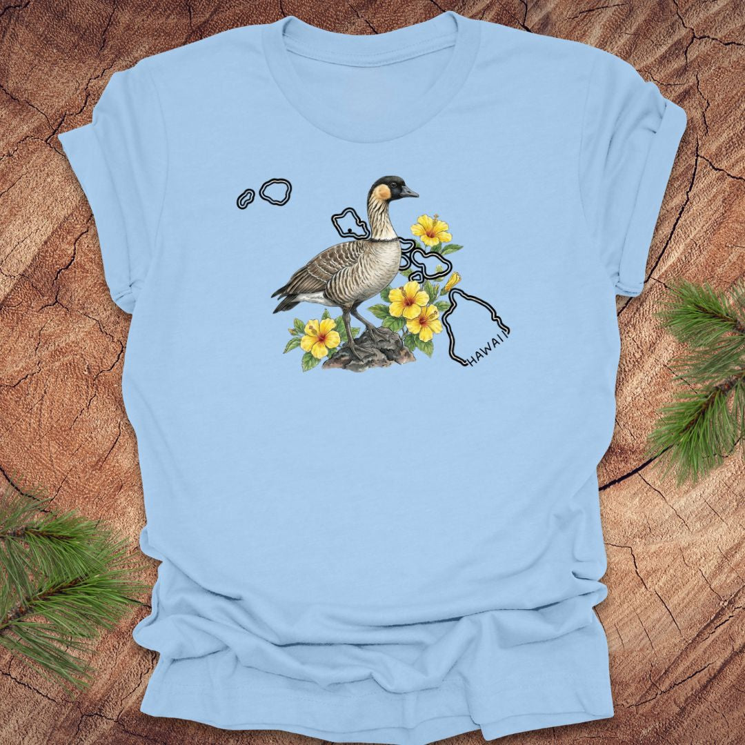 Baby blue t-shirt with a nene goose and hibiscus design over the outline of the state of Hawaii on a wooden surface