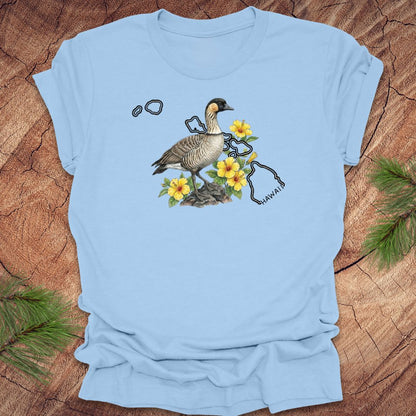 Baby blue t-shirt with a nene goose and hibiscus design over the outline of the state of Hawaii on a wooden surface