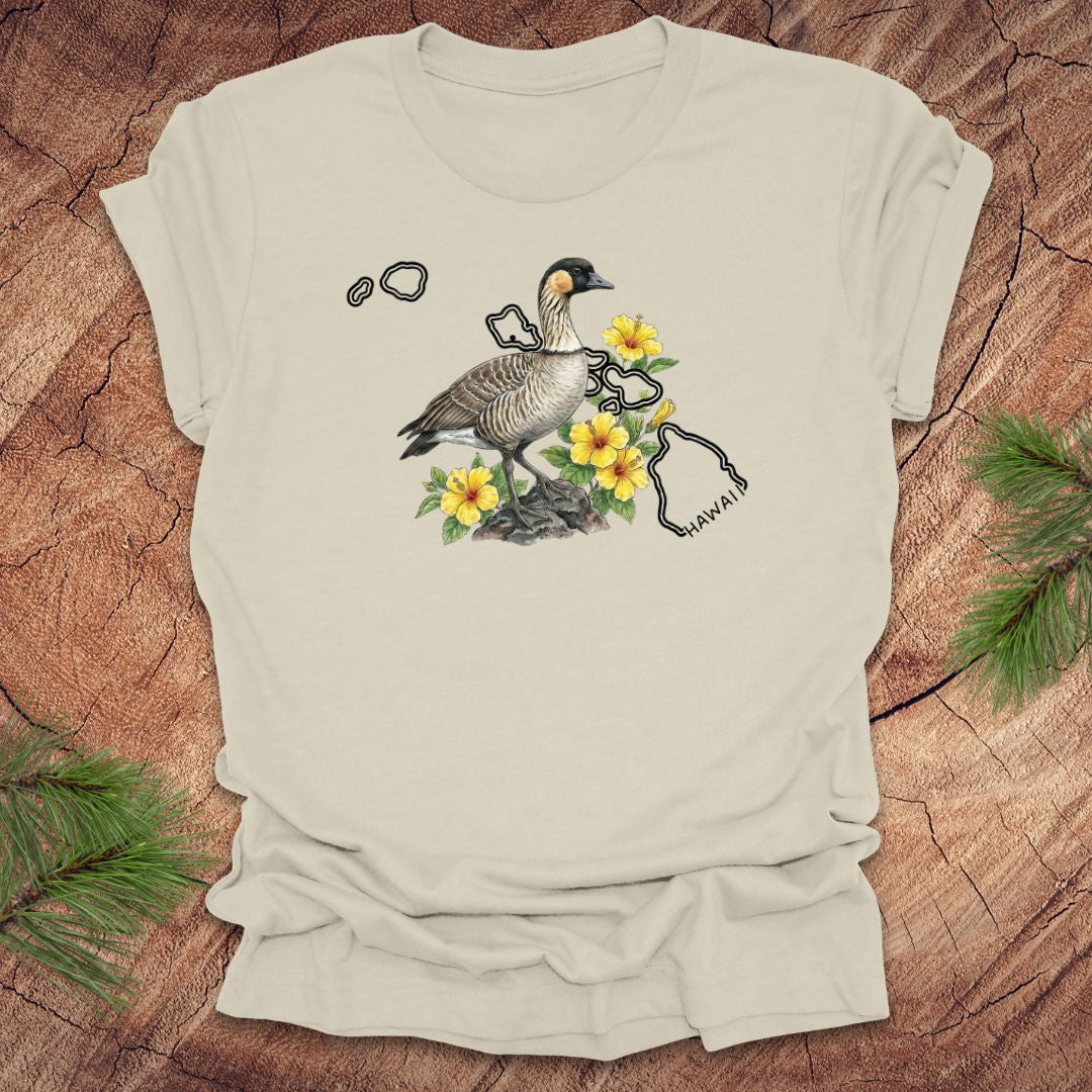 Beige t-shirt with a nene goose and hibiscus design over the outline of the state of Hawaii on a wooden surface