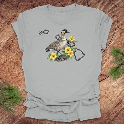 Silver t-shirt with a nene goose and hibiscus design over the outline of the state of Hawaii on a wooden surface