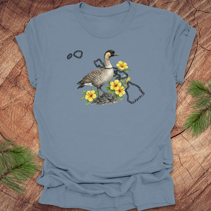 Steel blue t-shirt with a nene goose and hibiscus design over the outline of the state of Hawaii on a wooden surface