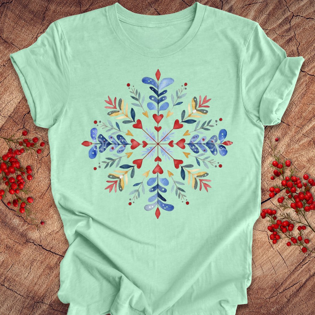A mint green short-sleeve t-shirt with a colorful watercolor snowflake design on the front, laid flat on a rustic wood background with red berries.