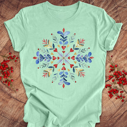 A mint green short-sleeve t-shirt with a colorful watercolor snowflake design on the front, laid flat on a rustic wood background with red berries.