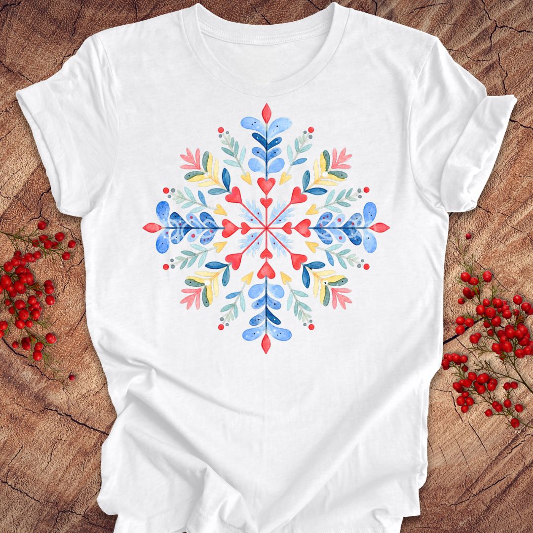 A white short-sleeve crewneck t-shirt featuring a colorful watercolor snowflake design on the front, styled flat on a rustic wood background with red berries.