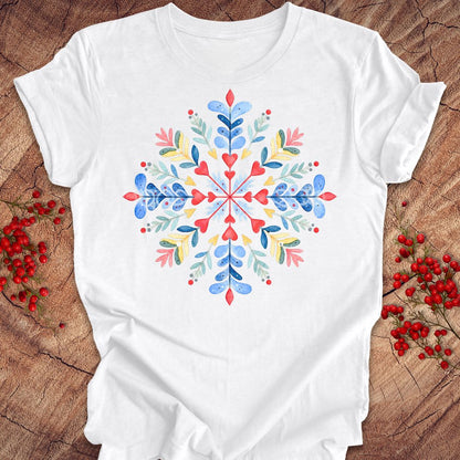 A white short-sleeve crewneck t-shirt featuring a colorful watercolor snowflake design on the front, styled flat on a rustic wood background with red berries.