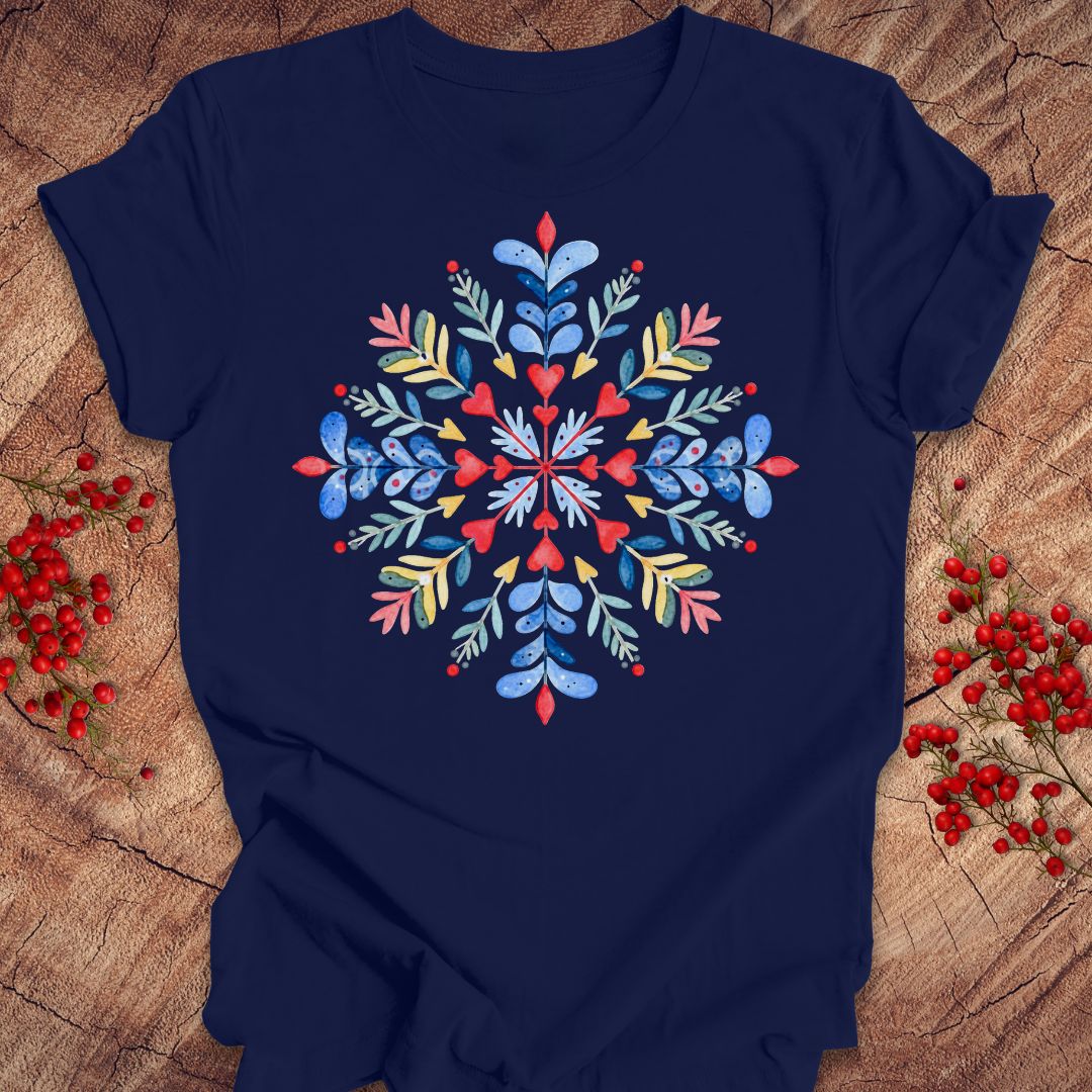 A navy blue short-sleeve t-shirt with a colorful, symmetrical floral snowflake design printed on the chest, styled flat on a rustic wood background with red berries.