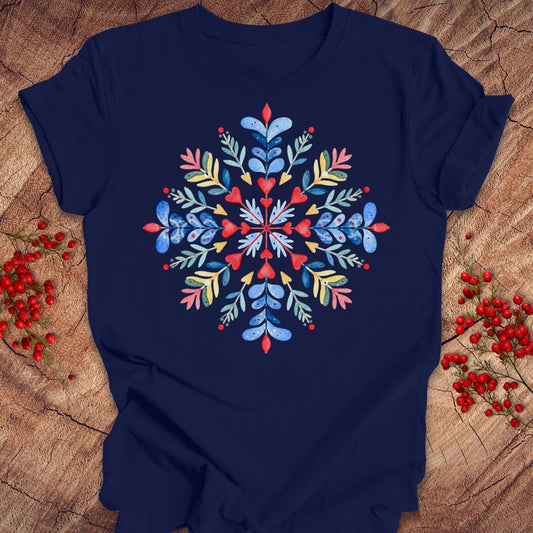 A navy blue short-sleeve t-shirt with a colorful, symmetrical floral snowflake design printed on the chest, styled flat on a rustic wood background with red berries.