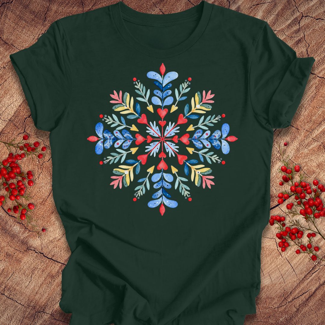 A dark forest green short-sleeve t-shirt with a colorful, symmetrical floral snowflake graphic on the chest, laid flat on a rustic wood background decorated with red berries.