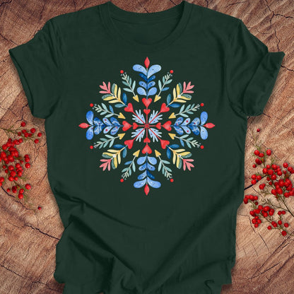 A dark forest green short-sleeve t-shirt with a colorful, symmetrical floral snowflake graphic on the chest, laid flat on a rustic wood background decorated with red berries.
