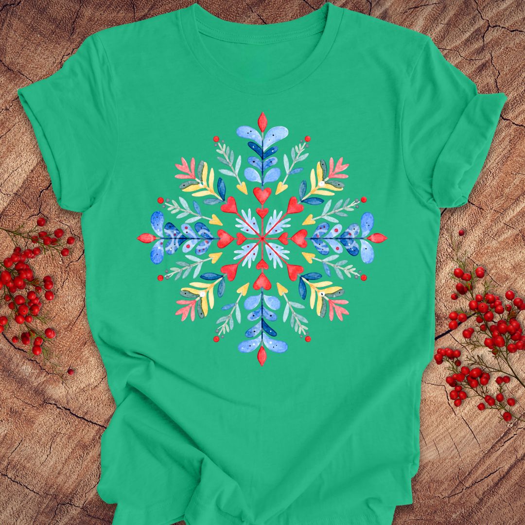 A green short-sleeve crewneck t-shirt with a colorful, symmetrical snowflake design printed on the chest, styled flat on a rustic wood background with red berries.
