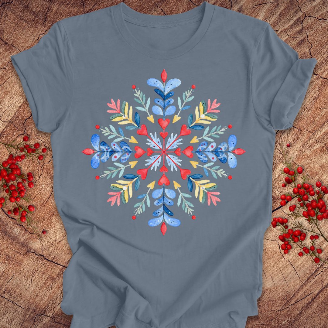 A slate blue short-sleeve t-shirt with a colorful watercolor snowflake graphic on the chest, styled flat on a rustic wood background with red berries.