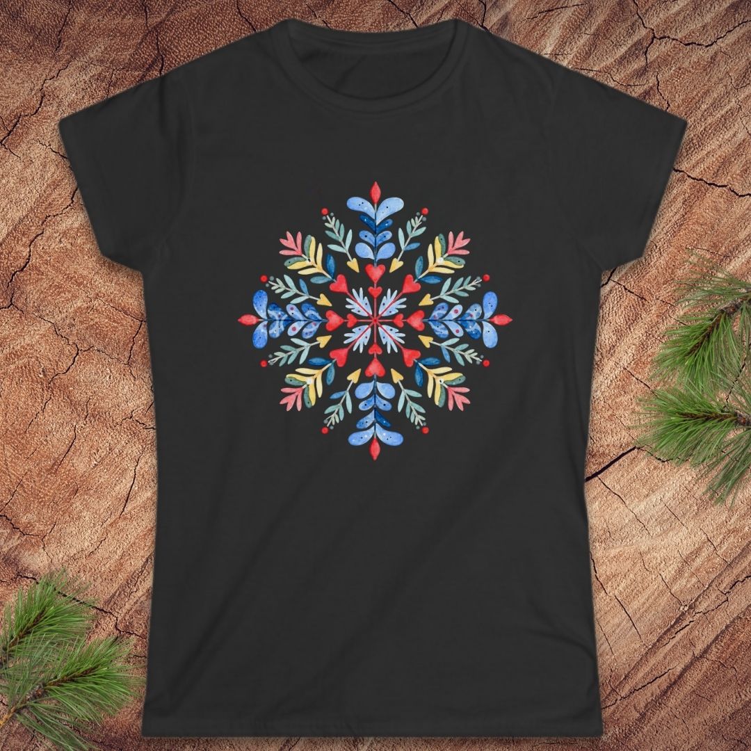 A black short-sleeve crewneck t-shirt featuring a colorful, symmetrical floral mandala graphic on the chest, laid flat on a textured wood background with pine needles in the corners.