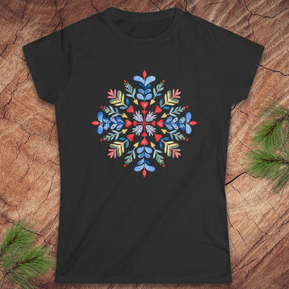 A black short-sleeve crewneck t-shirt featuring a colorful, symmetrical floral mandala graphic on the chest, laid flat on a textured wood background with pine needles in the corners.