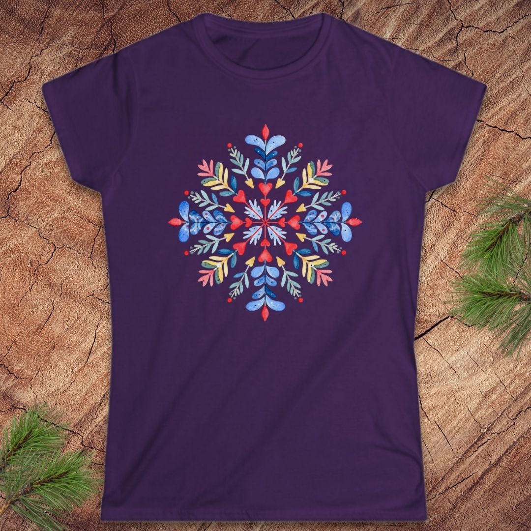 A purple short-sleeve crewneck t-shirt featuring a colorful, symmetrical mandala-style snowflake graphic on the chest, laid flat on a rustic wood background with pine needles in the corners.