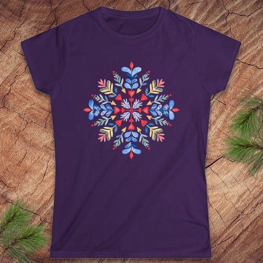 A purple short-sleeve crewneck t-shirt featuring a colorful, symmetrical mandala-style snowflake graphic on the chest, laid flat on a rustic wood background with pine needles in the corners.