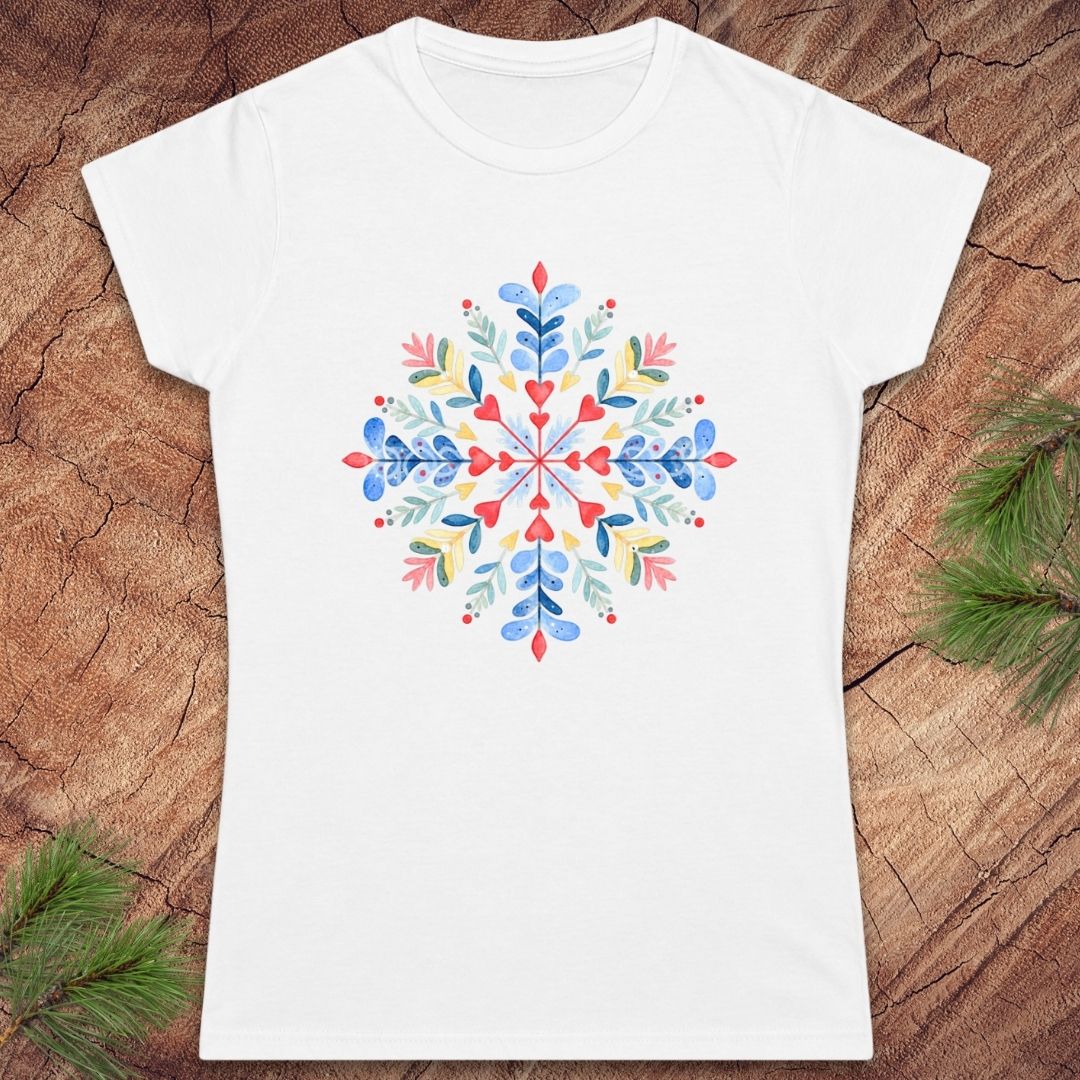A white short-sleeve crewneck t-shirt featuring a colorful, symmetrical watercolor snowflake design on the chest. The shirt is displayed flat on a rustic wood background with pine branches in the corners.
